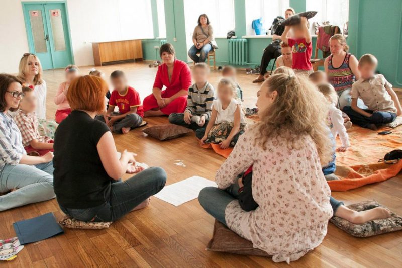 Каля 20 дзяцей сядзяць на падлозе ў колах вакол адной жанчыны, сярод іх ёсць дарослыя