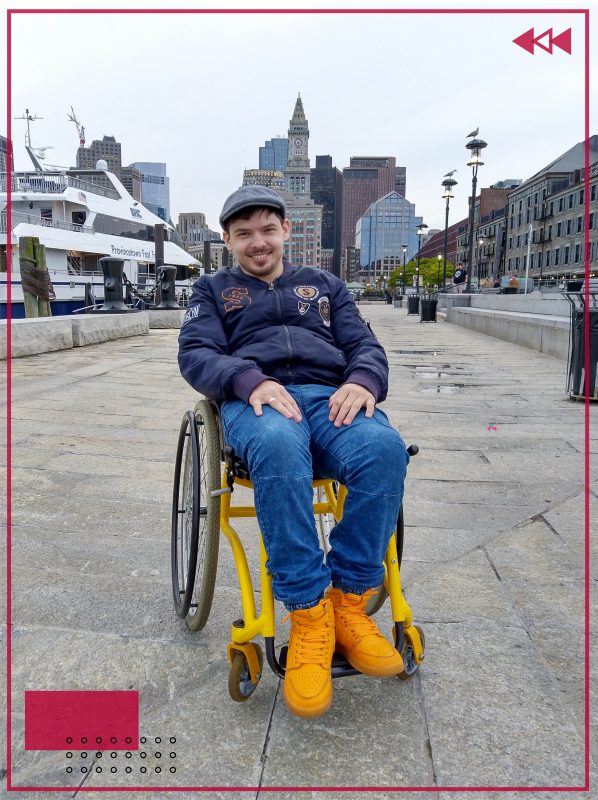 Герой материала сидит на коляске и улыбается на фоне яхты и небоскрёбов в Бостоне